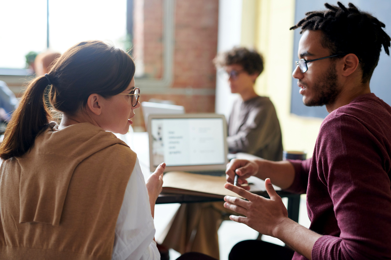 Two people are sitting at a desk, engaged in conversation. A laptop is open in front of them, while two other people work in the blurred background. The setting appears to be a modern, casual office or classroom.