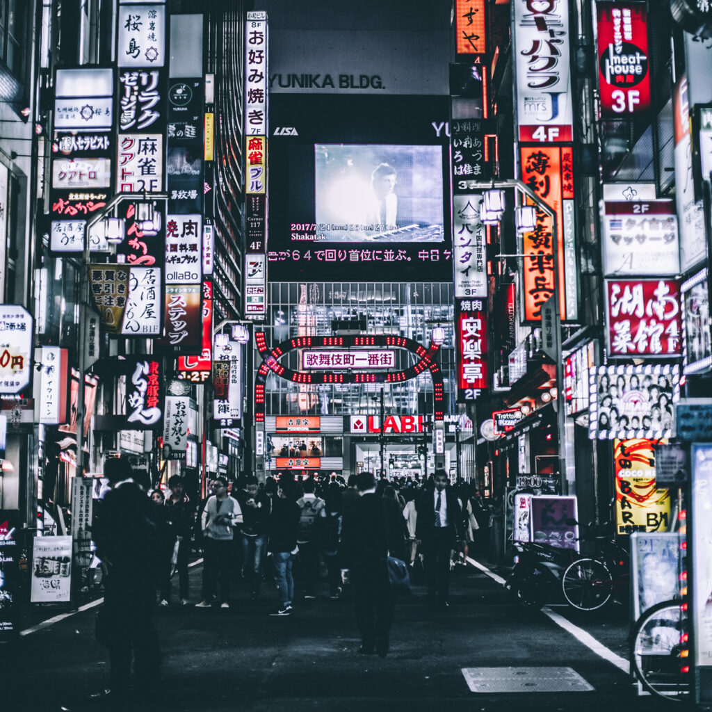 A busy street in Tokyo at night is filled with people and bicycles, surrounded by bright neon signs and advertisements in Japanese, with a large screen displaying a person in the background.