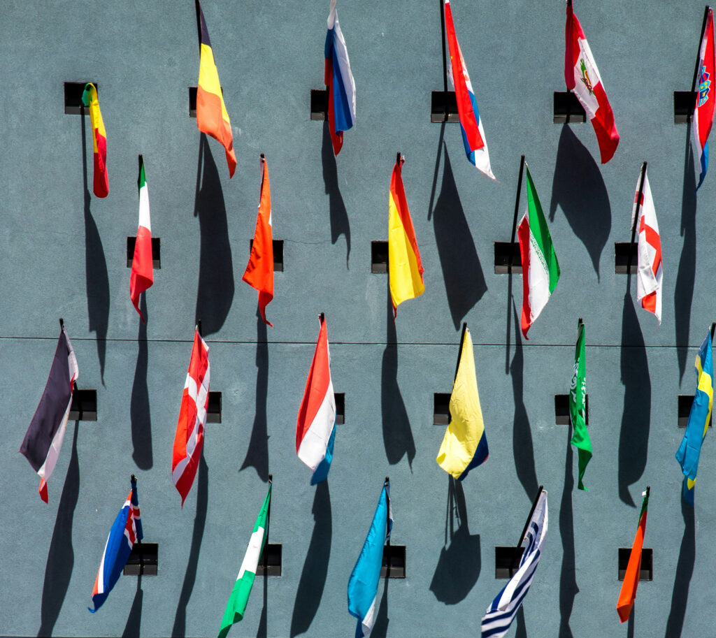 A row of various international flags hanging vertically on a blue wall, each casting a distinct shadow below due to bright sunlight. The wall has evenly spaced square fixtures holding the flagpoles.