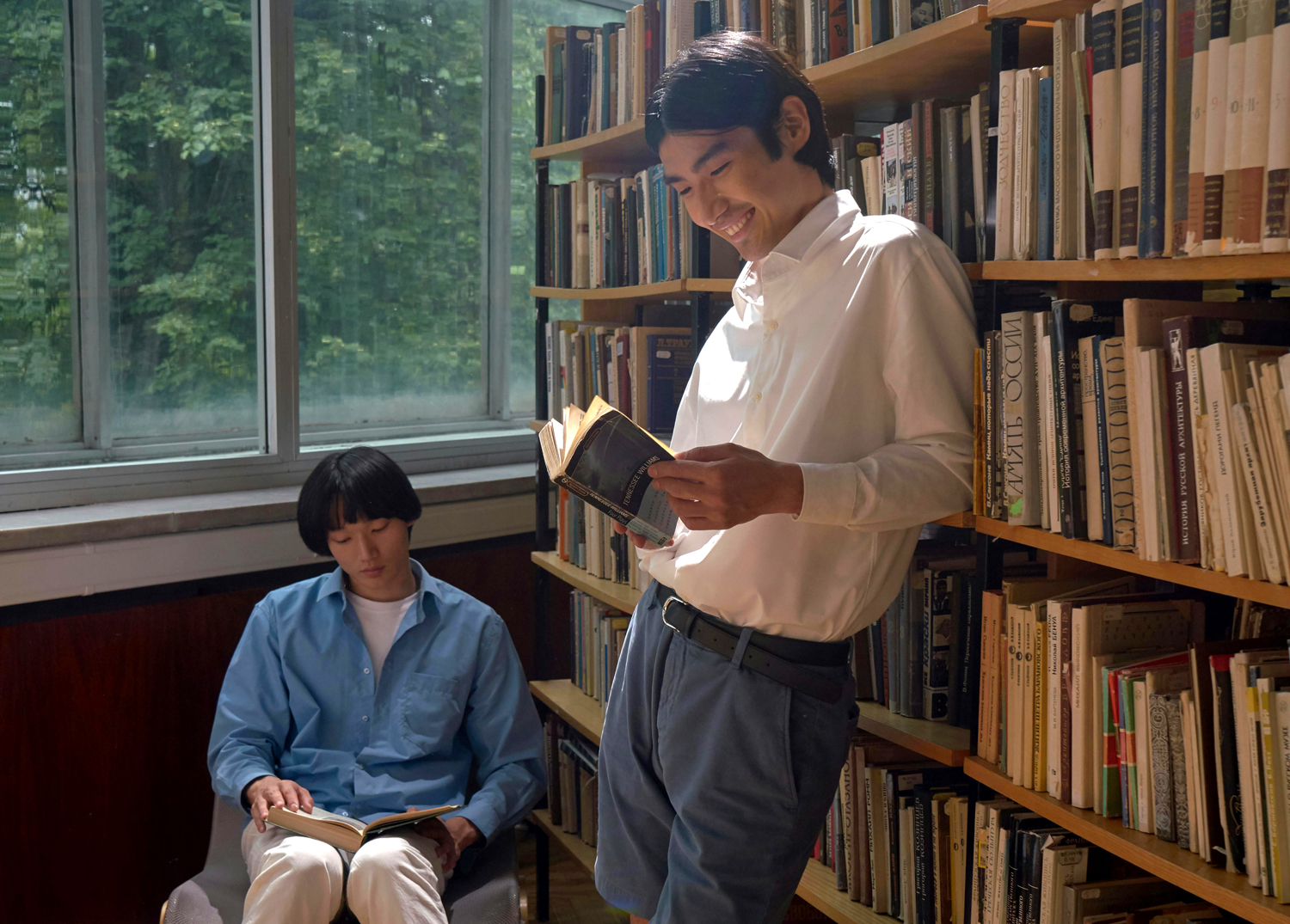 Two people in a library, one sitting and reading by a large window, and the other standing and smiling while reading a book. Shelves of books line the walls and sunlight filters through the window.