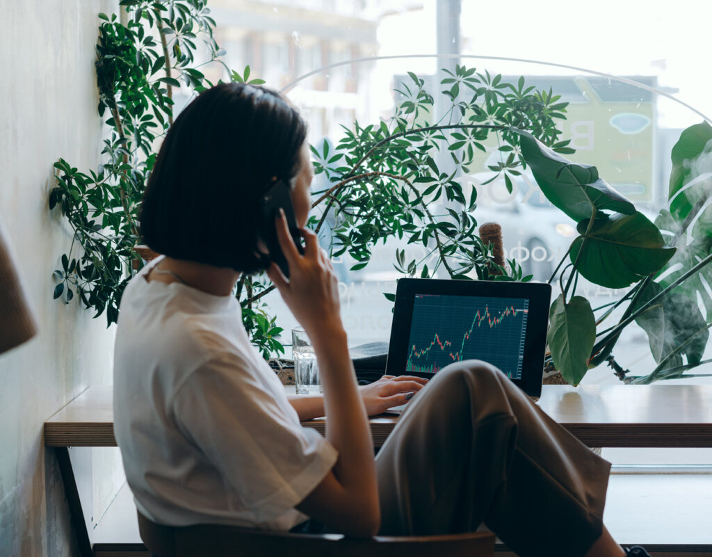 A woman sits at a desk by a window, talking on the phone and looking at a laptop displaying a line graph. There are green plants on the desk and natural light coming in through the window.
