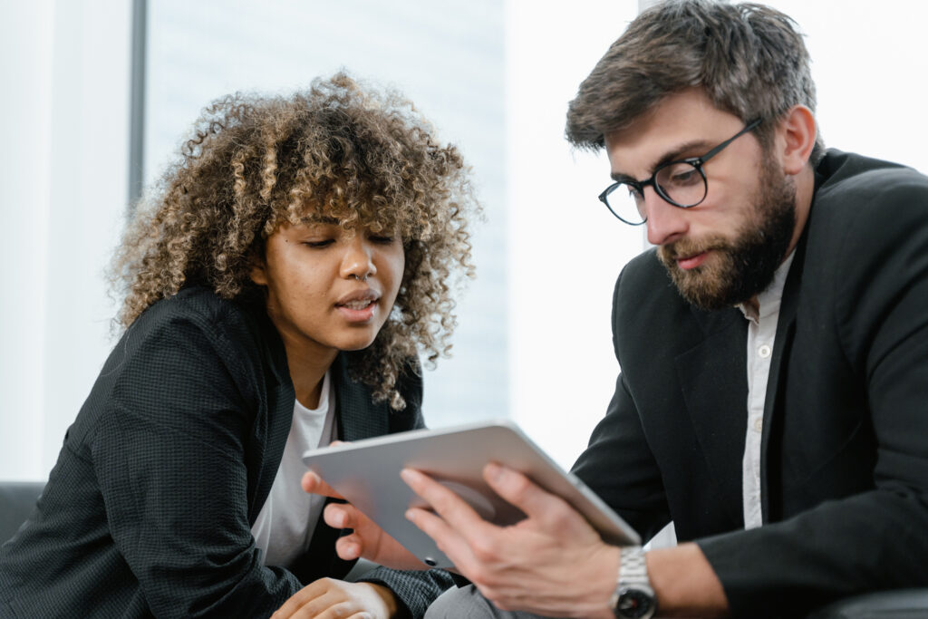 Two people in business attire sit closely together, looking intently at a digital tablet that one of them is holding, appearing to discuss or review content displayed on the screen.