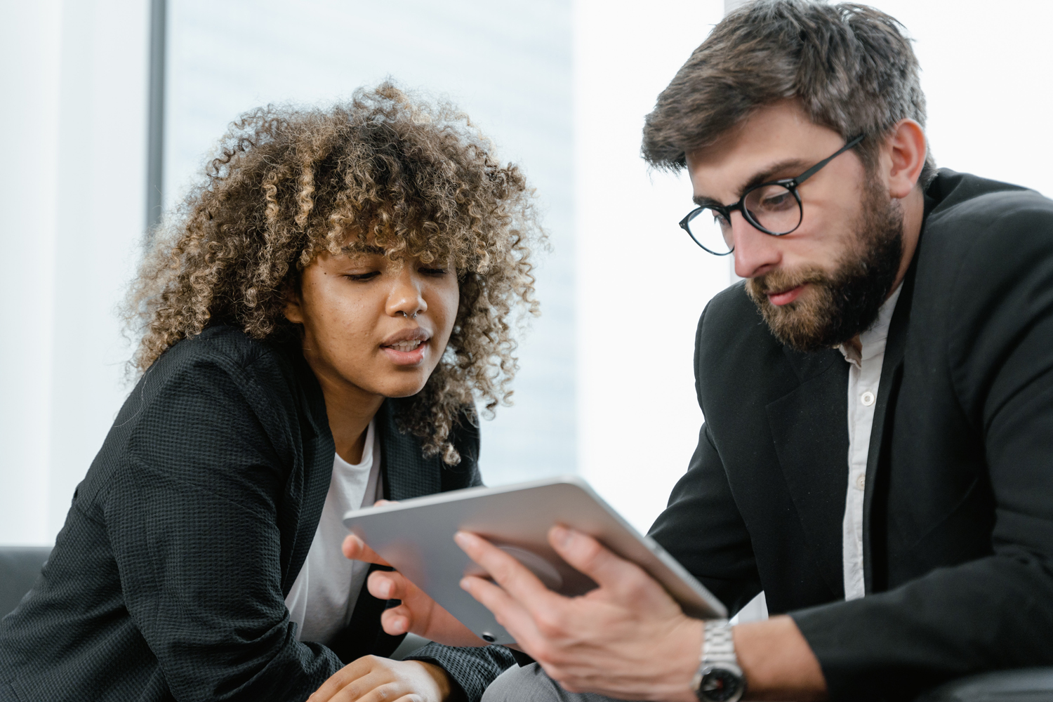 Two people in business attire sit closely together, looking intently at a digital tablet that one of them is holding, appearing to discuss or review content displayed on the screen.