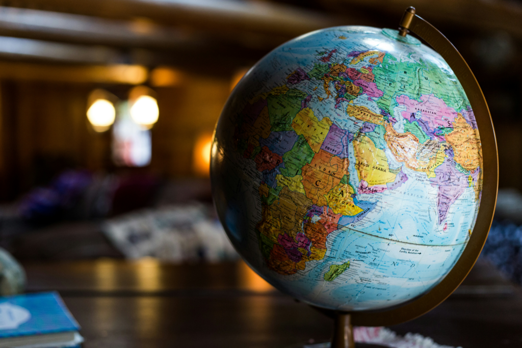 A colorful globe sits on a wooden surface indoors, displaying parts of Europe, Africa, and Asia. The background is softly blurred with warm lighting and indistinct objects.