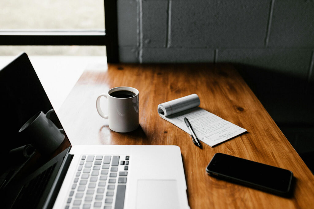 A wooden desk with an open laptop, a white coffee mug, a notepad with handwritten notes, a black pen, and a smartphone, next to a window letting in natural light.