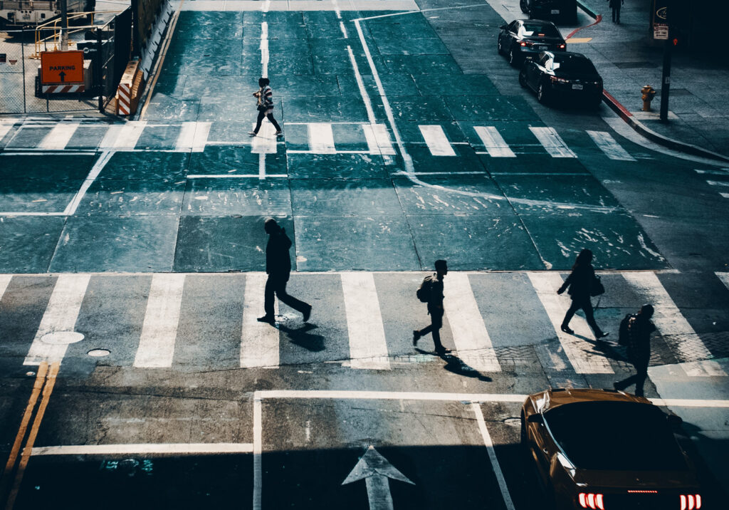 An overhead view of a city intersection shows several people walking across crosswalks, with cars waiting at the corners and a yellow car in the foreground. The scene is partially shadowed by buildings.