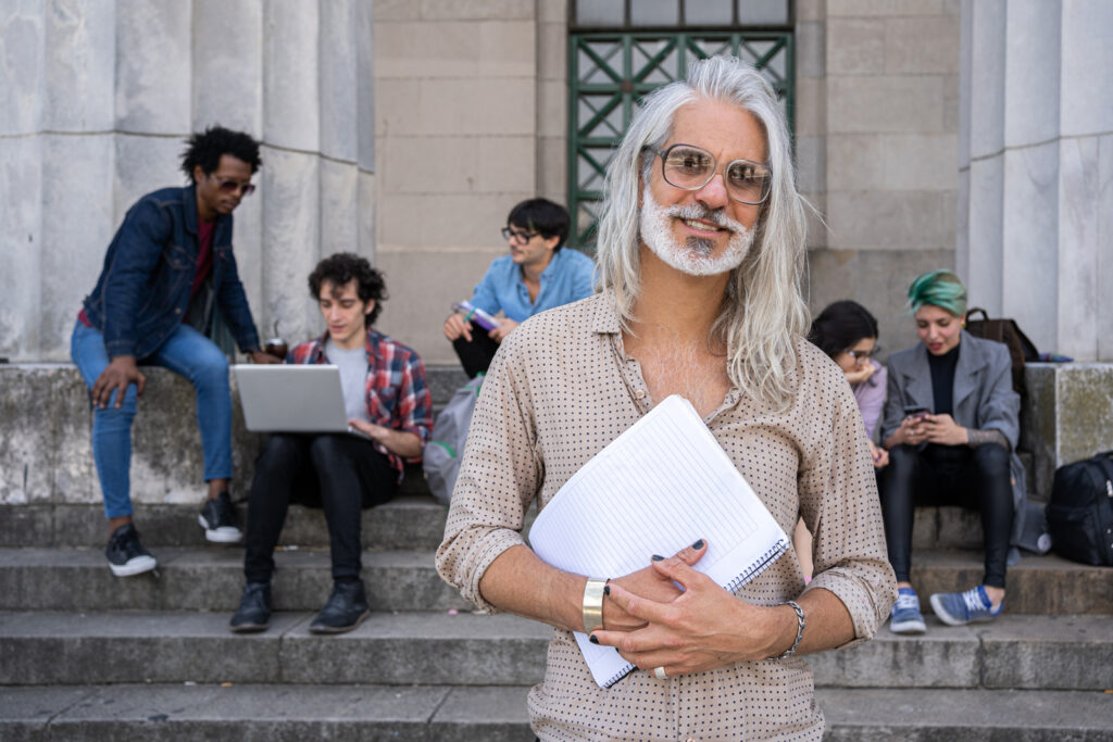 A person with long gray hair and glasses holds notebooks and smiles, standing in front of stone steps where a group of five people sits, talking, using a laptop, and looking at phones.