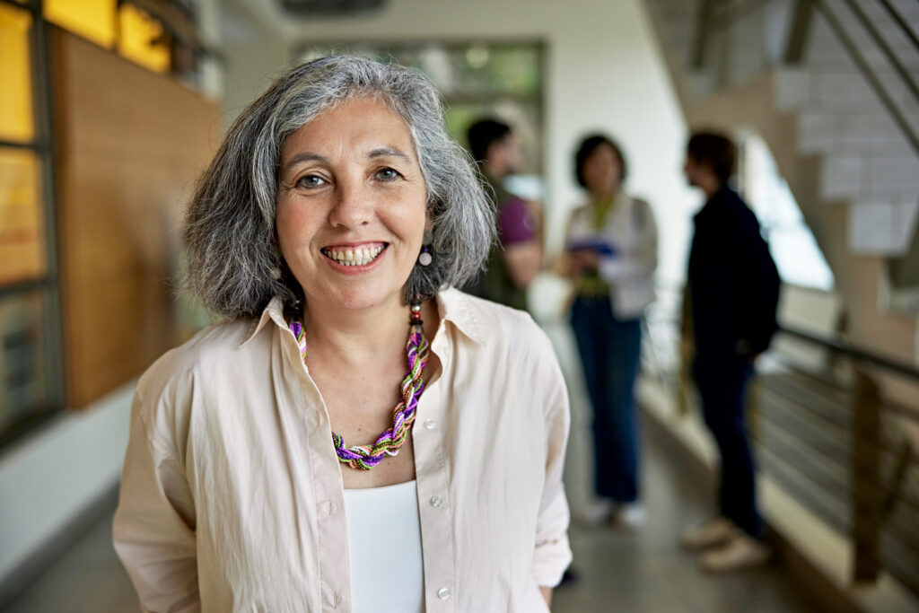 A smiling woman with gray hair and a colorful necklace stands in focus in a hallway, while three people chat in the blurred background near a staircase.