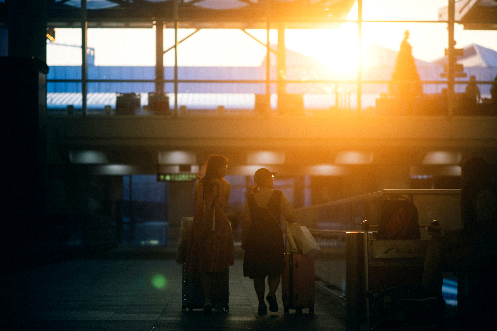 Two people walk side by side through an airport terminal at sunset, each pulling a suitcase, with warm sunlight streaming through large windows in the background.