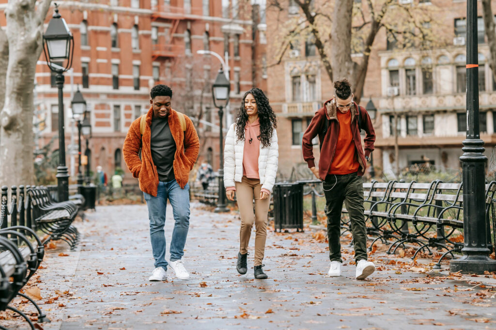 Three young adults walk together and chat in a city park on a fall day, surrounded by benches and trees with fallen leaves. They look happy and relaxed.