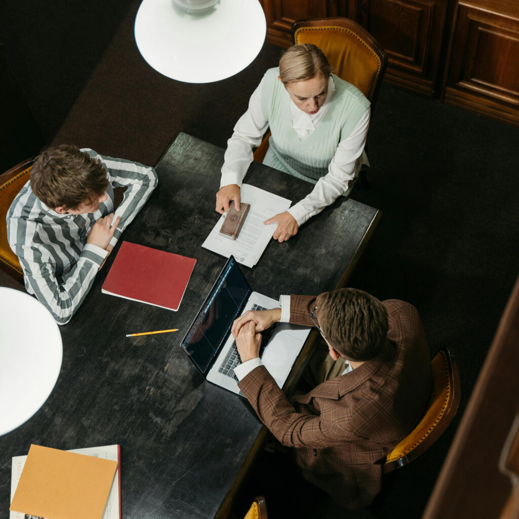 Three people sit around a dark wooden table with documents, a laptop, and notebooks, engaged in discussion. One person points at a paper. The scene is viewed from above under two hanging lights.