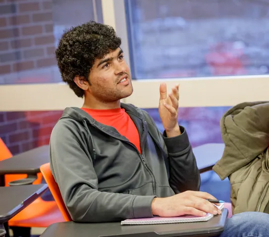 A student with curly hair sits at a desk in a classroom, holding a pen and notebook, and gestures with one hand as if asking a question or making a point.
