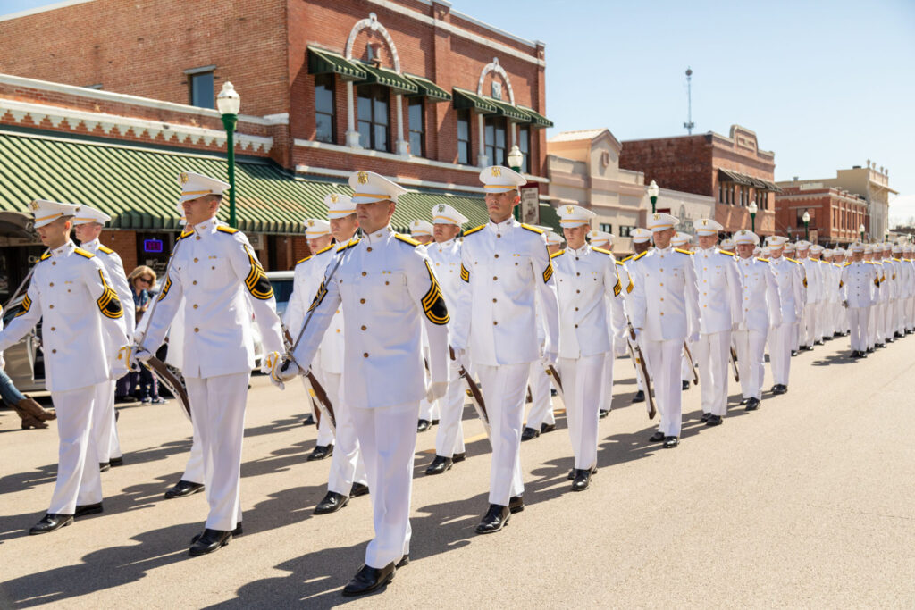 A group of uniformed cadets in white dress uniforms marches in formation down a sunny street, flanked by historic brick buildings. Spectators watch from the sidewalk.