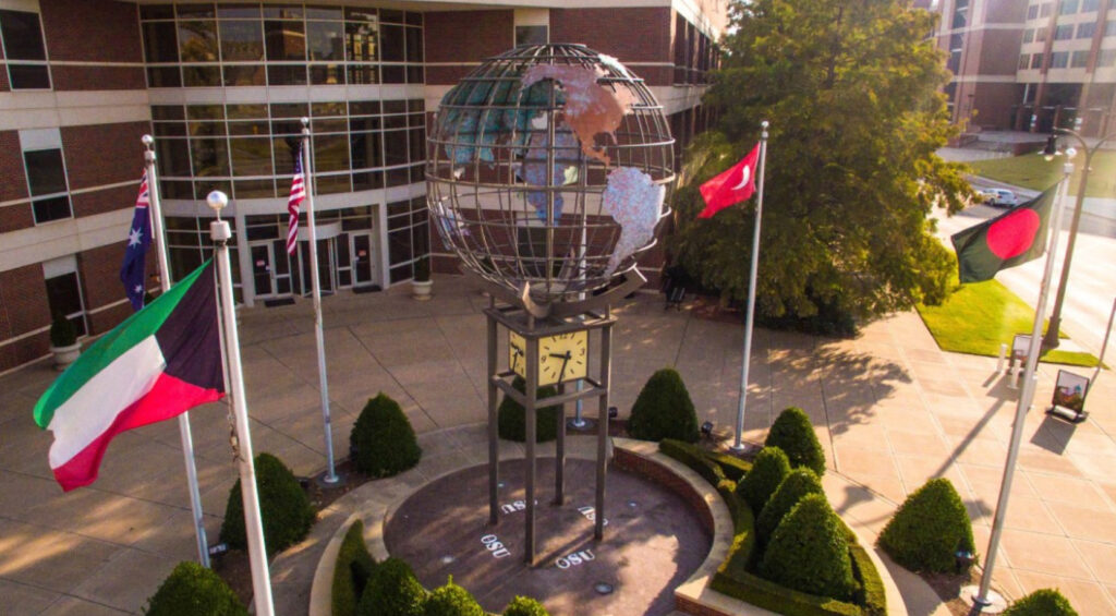 A large metal globe sculpture with a clock at its base stands in a plaza, surrounded by trimmed bushes and flagpoles displaying international flags in front of a modern brick building.