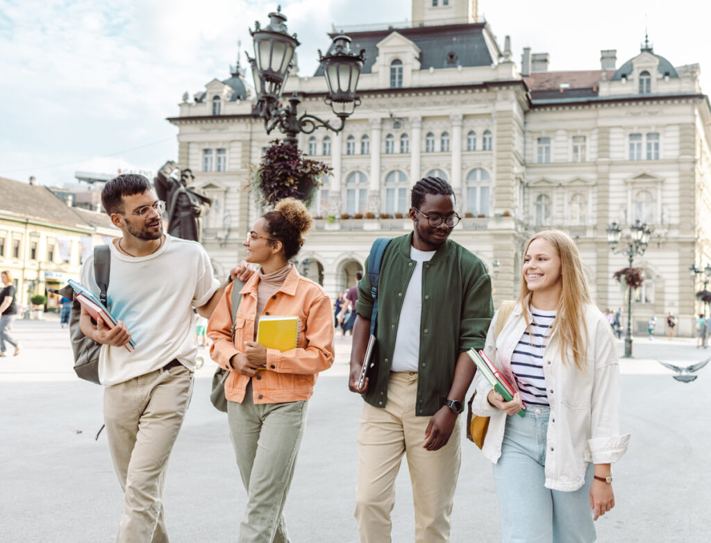Four young adults walk together outside, holding books and smiling in conversation. They are in front of a large, ornate building with arched windows and decorative lamps. It appears to be a university or historic site.