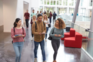 A group of college students walk and talk in a modern, bright hallway with large windows. Three students are in the front, holding books and backpacks, while more students walk behind them. Red chairs are visible on the right.