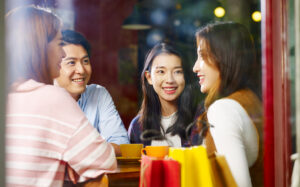 Four young adults sit at a café table, smiling and conversing. Two women and two men are visible through a window, with coffee cups and shopping bags on the table, creating a warm, friendly atmosphere.