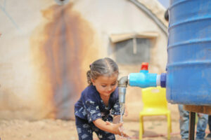 A young girl with braided hair smiles as she washes her hands under running water from a blue outdoor tap, with a yellow chair and a weathered building in the background.