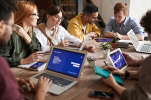 Six people sit around a wooden table with laptops, tablets, notebooks, and potted plants, collaborating and discussing work in a modern office setting.
