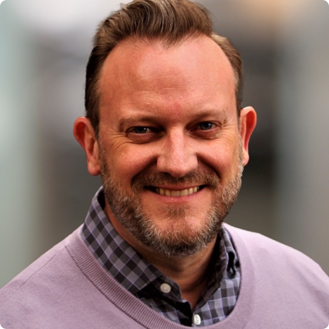 A man with short brown hair and a beard smiles at the camera. He is wearing a light purple sweater over a checkered shirt, and the background is softly blurred.