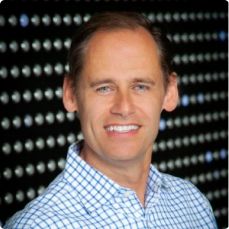 A smiling man with light skin and light brown hair, wearing a blue and white checkered shirt, stands in front of a background with evenly spaced small lights.
