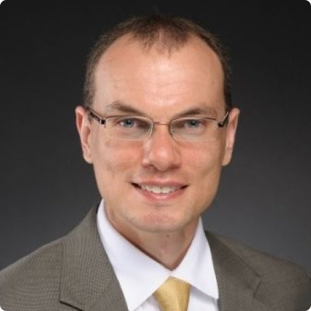 A man wearing glasses, a gray suit, a white shirt, and a yellow tie, smiling at the camera against a dark background.