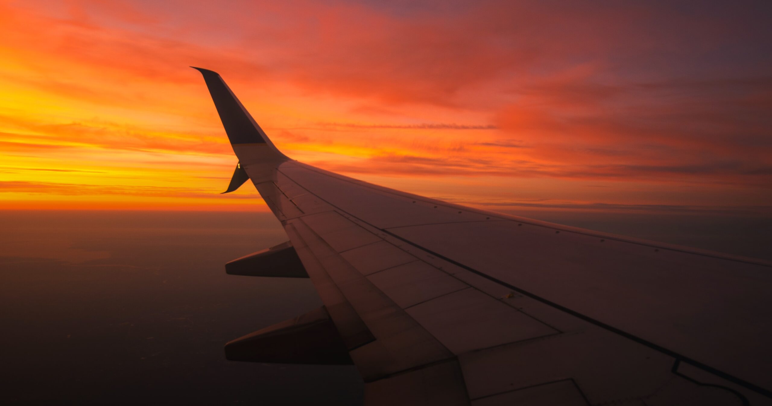 A view of an airplane wing in flight at sunset, with vibrant orange and yellow clouds filling the sky.
