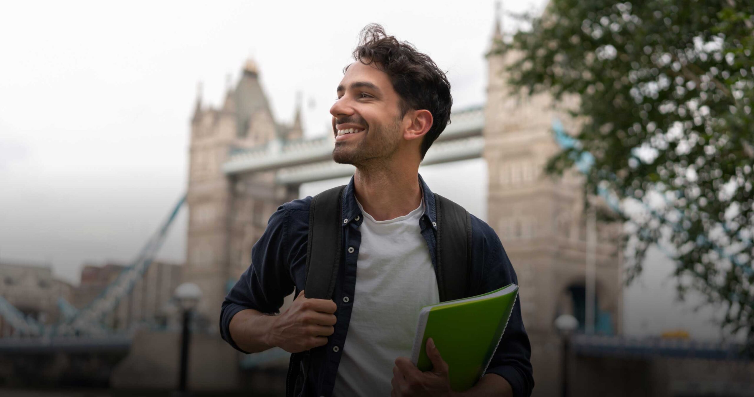 A young man with dark hair smiles while holding a green notebook and wearing a backpack. He stands outdoors in front of London’s Tower Bridge on a cloudy day, with trees visible on the right side of the image.
