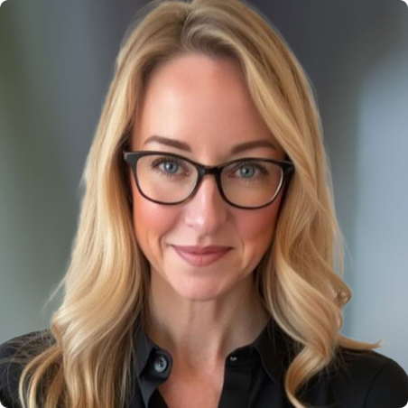 A woman with long blonde hair and glasses smiles softly. She is wearing a black blouse and stands against a blurred, neutral background.