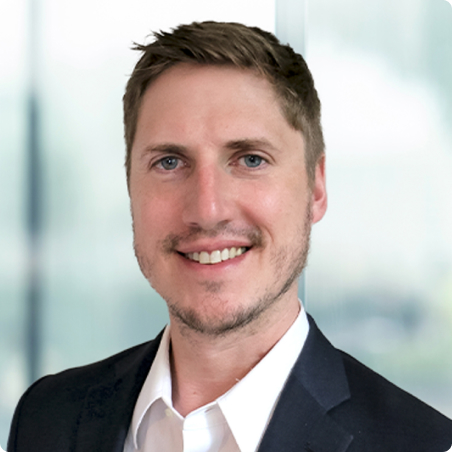 A man with short light brown hair, light facial stubble, and blue eyes is smiling. He is wearing a white shirt and a dark suit jacket, standing in front of a blurred office background.