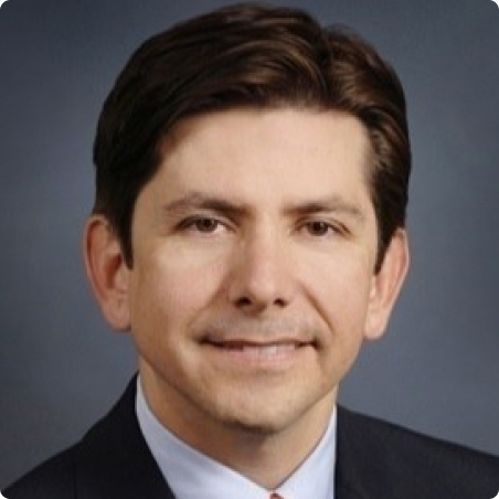 A man with short dark hair, wearing a dark suit, white shirt, and tie, smiles slightly in a professional portrait against a gray background.