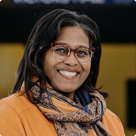 A woman with long dark hair and glasses smiles at the camera. She is wearing an orange coat and a patterned scarf, with a dark blurred background behind her.