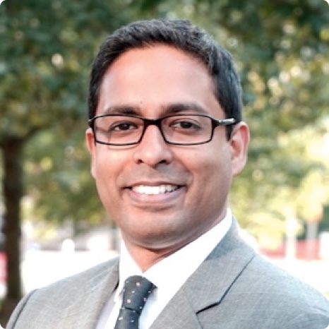 A man wearing glasses and a gray suit with a white shirt and black polka dot tie smiles outdoors with leafy green trees in the blurred background.