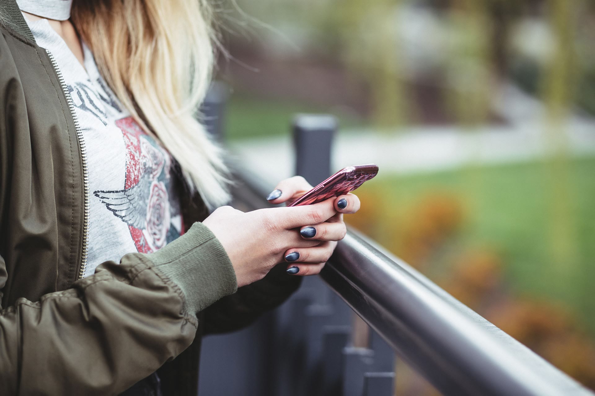 A person with long blonde hair, wearing a green jacket and graphic t-shirt, stands by a railing outdoors and uses a pink smartphone. The background is blurred with greenery visible.