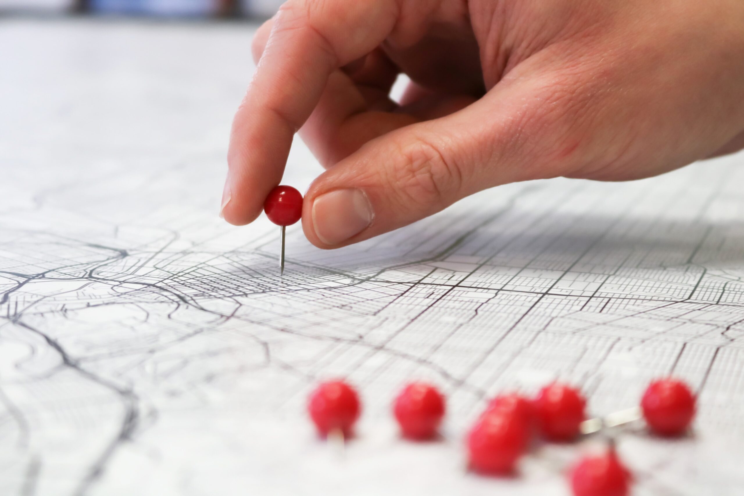 A hand places a red pushpin on a map, with several other red pushpins lying nearby on the map’s surface. The map is detailed with streets and lines.