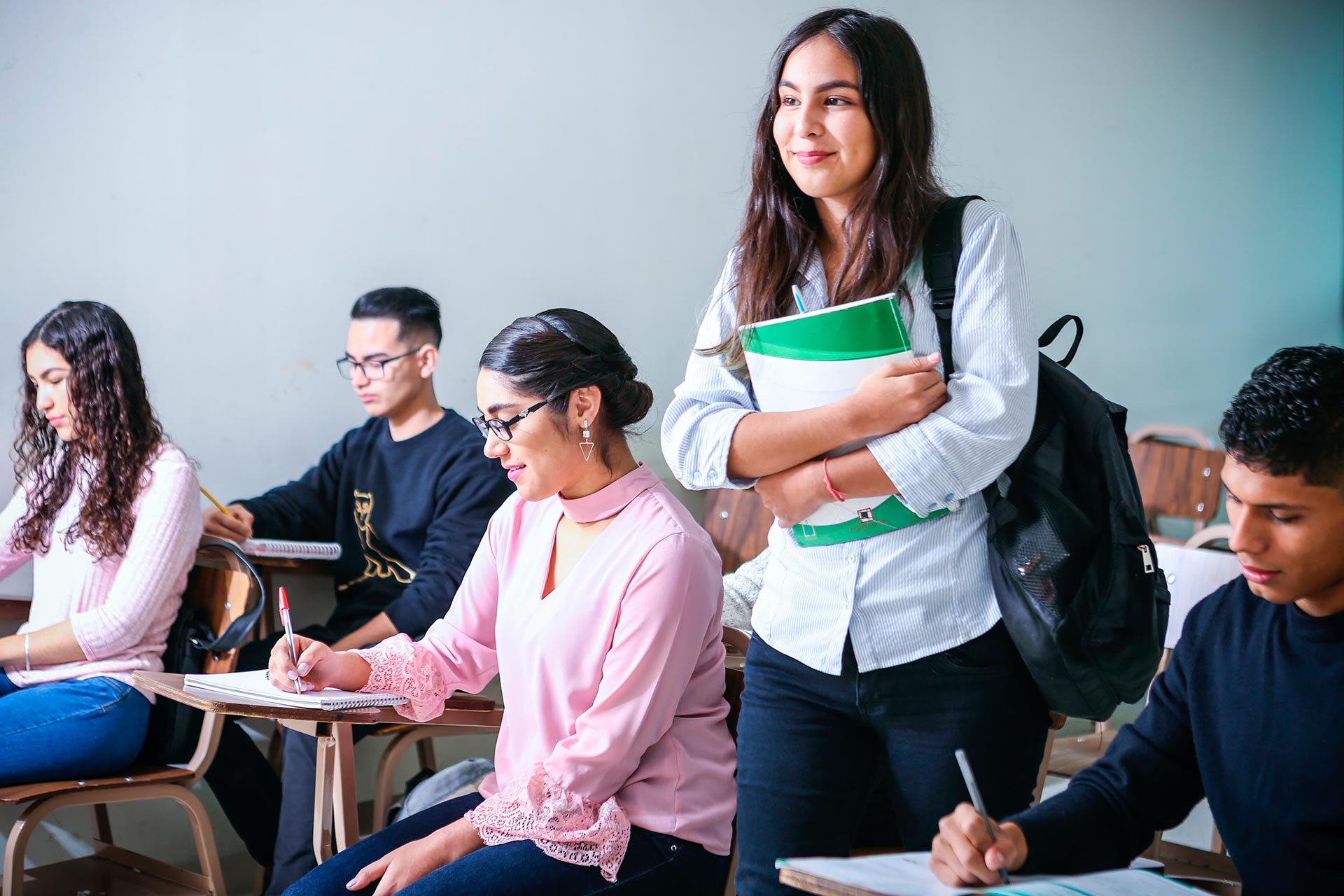 A smiling student stands holding books and a backpack in a classroom, while four other students sit at desks, writing and focusing on their work.