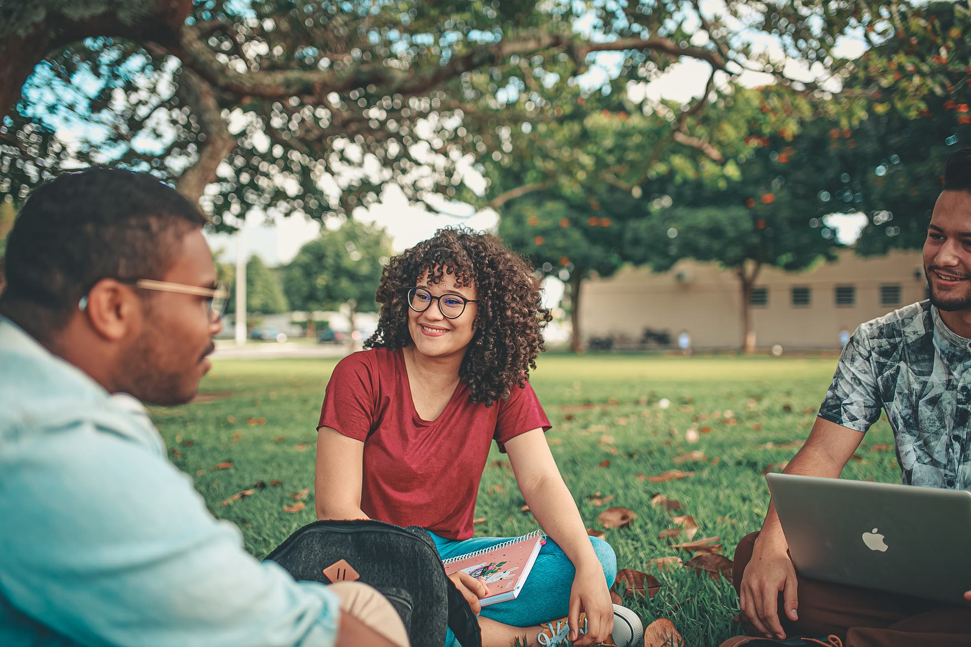 Three young adults sit on grass under a large tree, talking and smiling. One person holds a notebook, another has a laptop. Theyre surrounded by greenery on a sunny day, suggesting a casual outdoor study or hangout session.