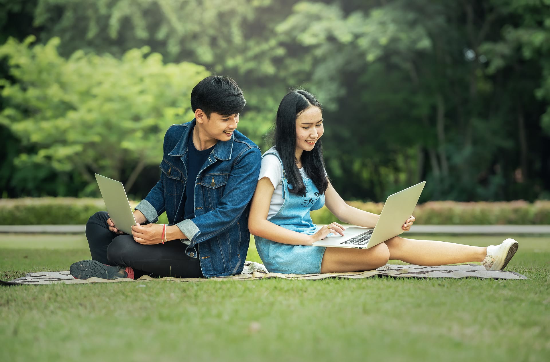 Two young people sit back-to-back on a blanket outdoors, each using a laptop. They appear to be studying or working, with green trees and grass in the background. Both are smiling and look focused.