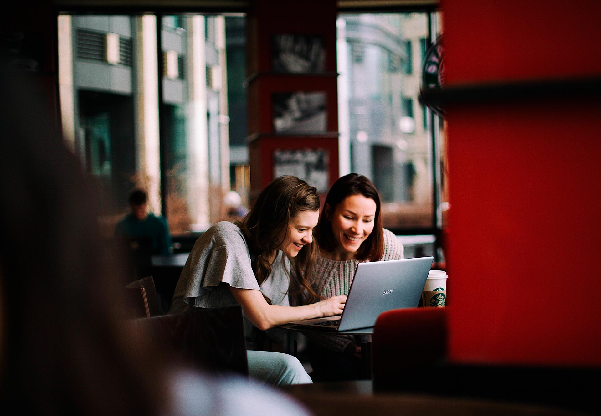 Two women sitting at a table in a cafe smiling and looking at a laptop screen together, with bright natural light coming through large windows in the background.