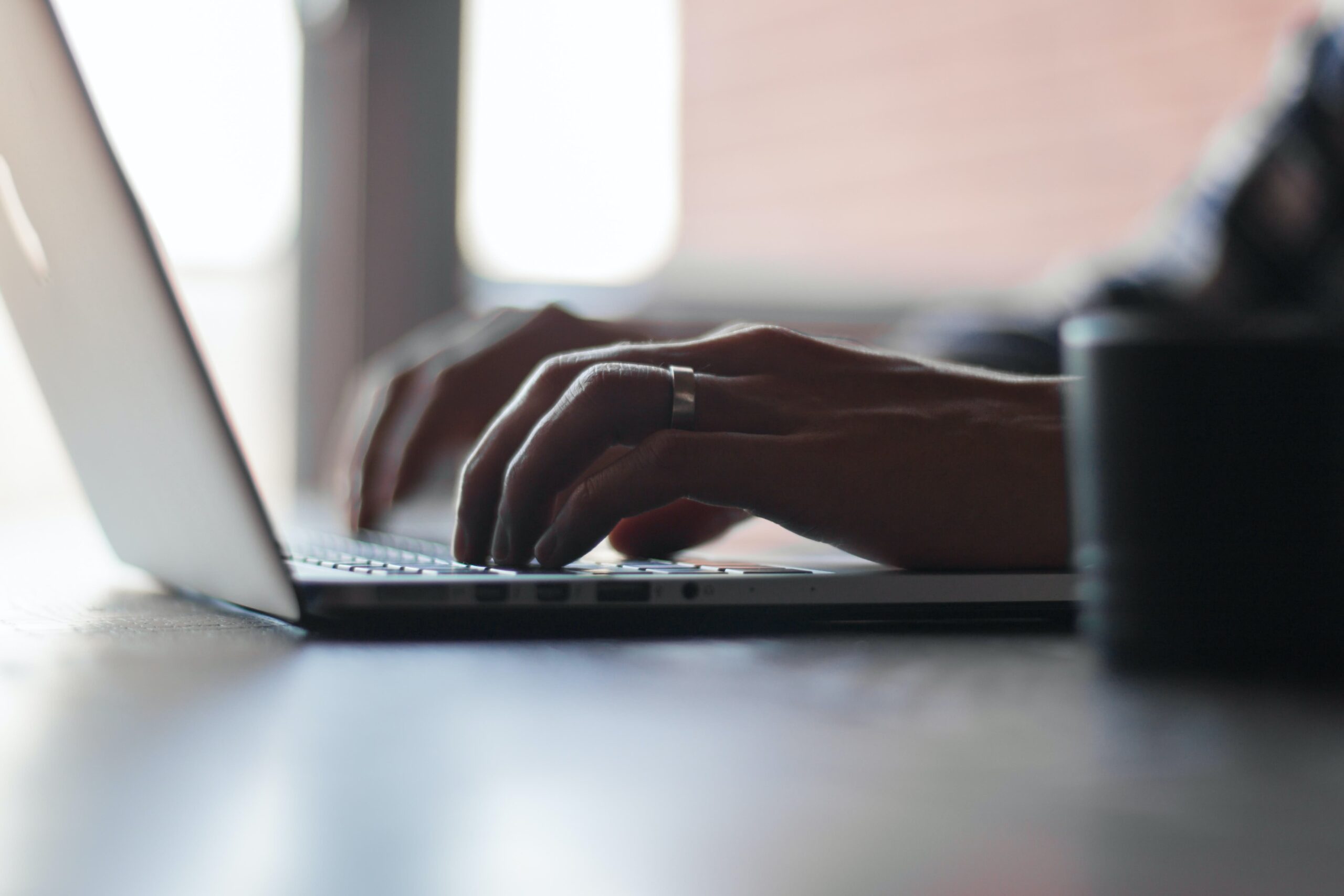 Close-up of a persons hands typing on a laptop keyboard, with soft natural light coming from a nearby window. The person is wearing a ring on one finger. The background is blurred.