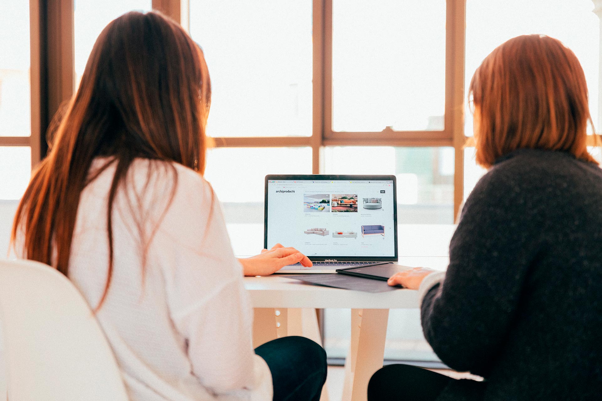 Two people sit at a table facing a laptop, looking at a website together. Sunlight streams through large windows in the background, creating a bright work environment.