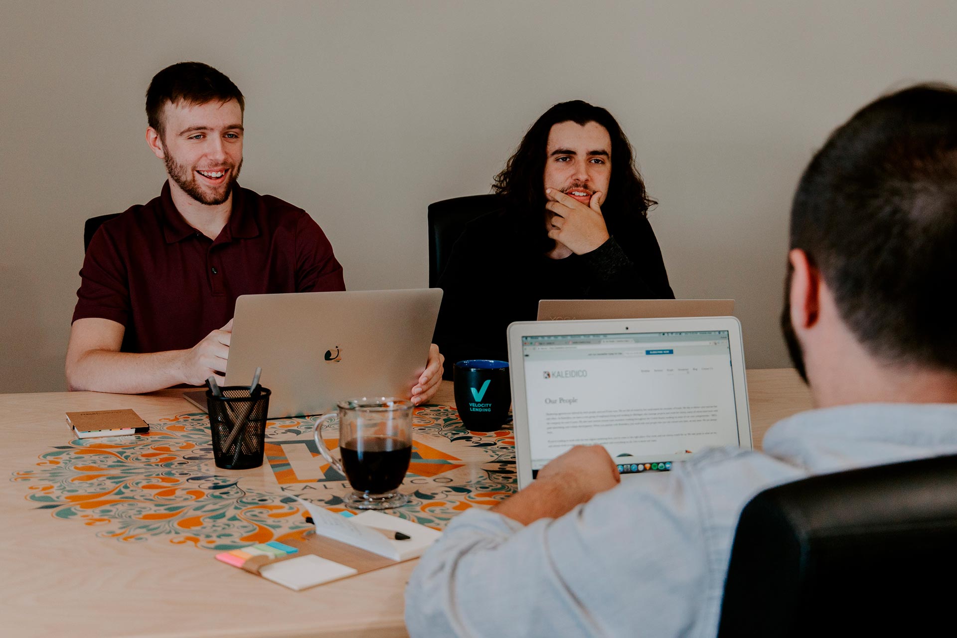Three people sit around a table with laptops, engaged in a meeting or discussion. Two face forward, smiling and attentive, while the third is seen from behind, reading from a laptop. Office supplies and coffee cups are on the table.