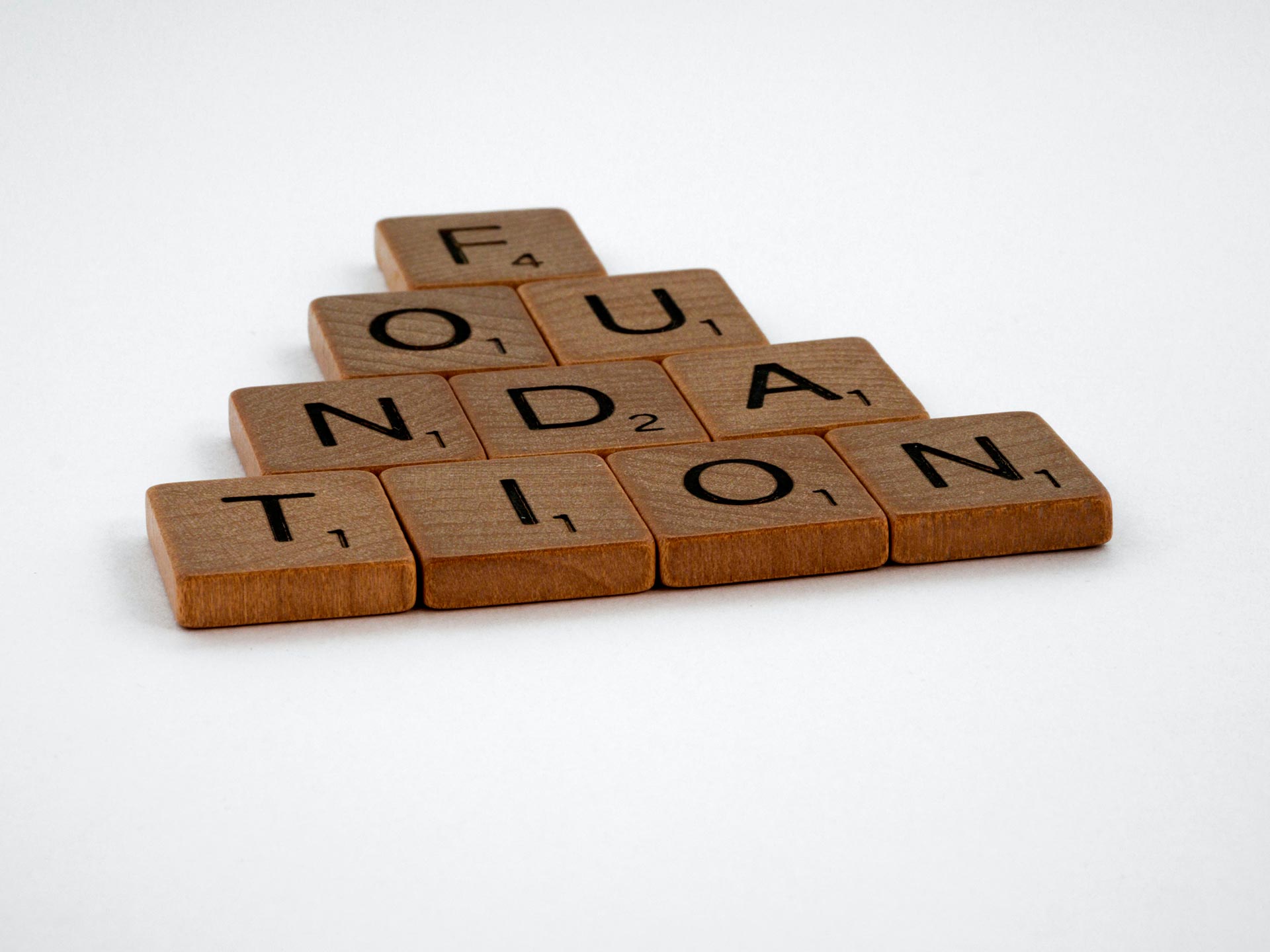 Pyramid-shaped arrangement of Scrabble tiles spelling the word FOUNDATION on a white background.