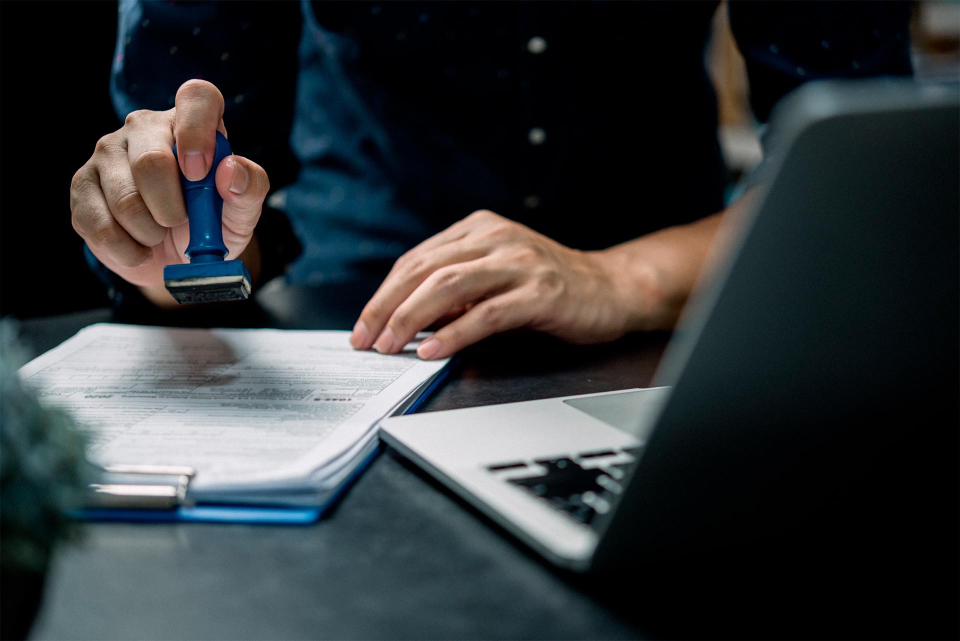 A person stamps a document on a clipboard while sitting at a desk next to an open laptop.