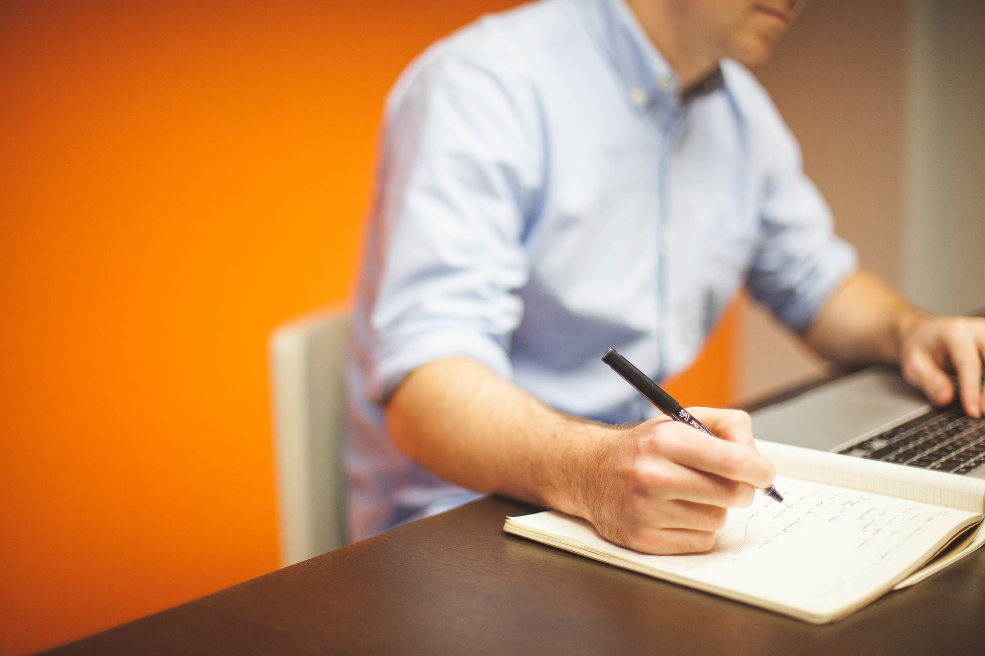 A person in a light blue shirt writes in a notebook with one hand while using a laptop with the other, seated at a desk in front of an orange wall.