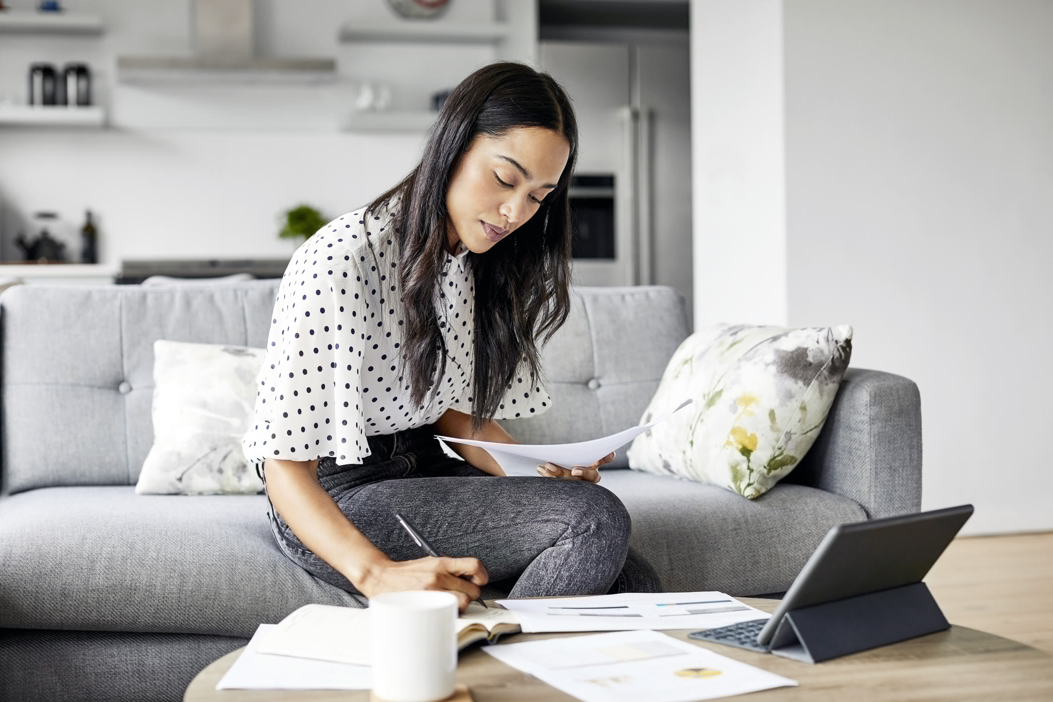 A woman sits on a gray sofa at a coffee table, working with papers, a notebook, and a tablet. She is writing, focused, with a mug nearby. The setting appears to be a modern, bright living room.