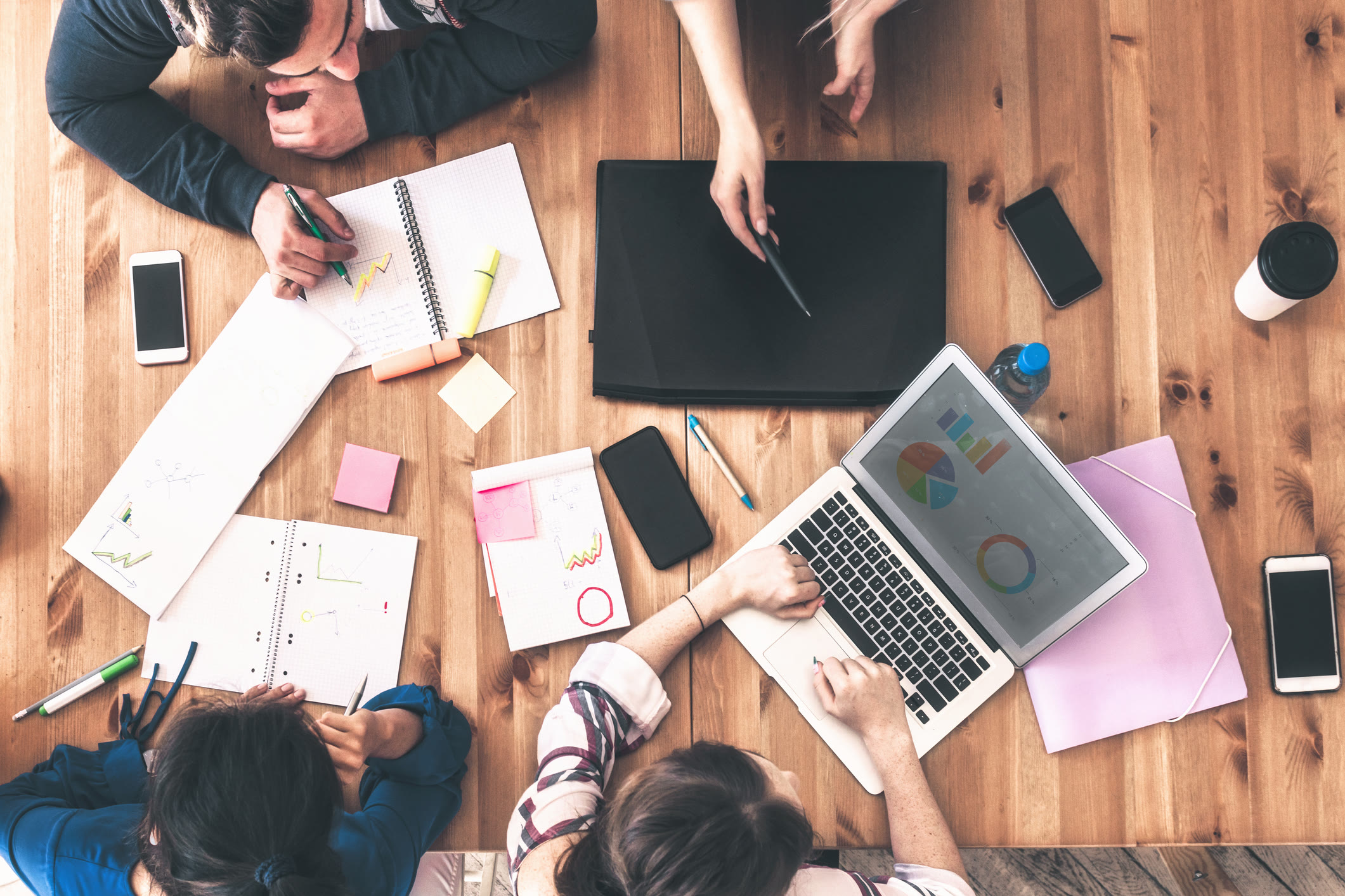 Four people sit around a table with a laptop showing colorful charts, notebooks, pens, papers, and smartphones, actively collaborating and working together on a project.