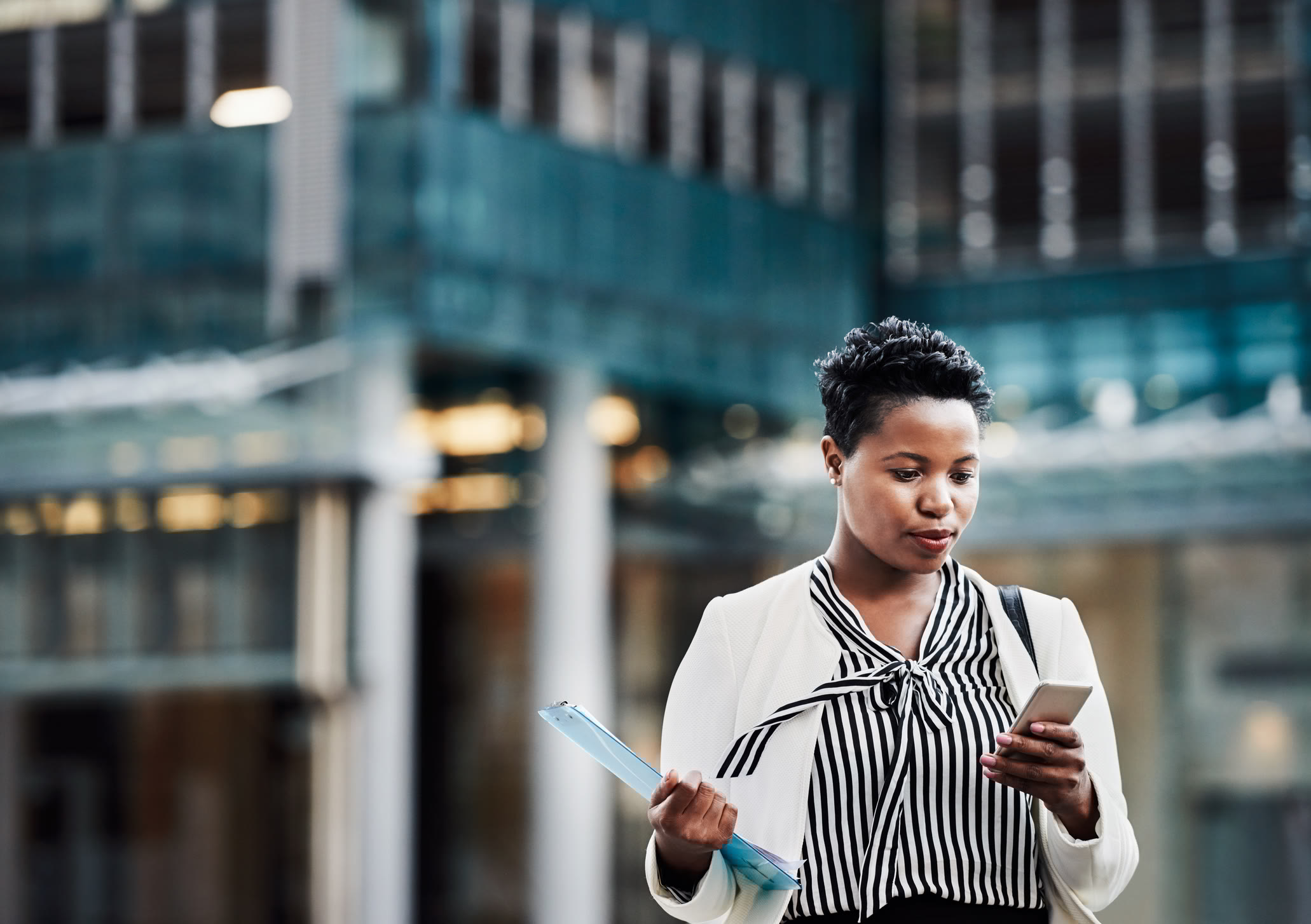 A woman in business attire stands outside an office building, holding documents in one hand and looking at her smartphone with a focused expression.