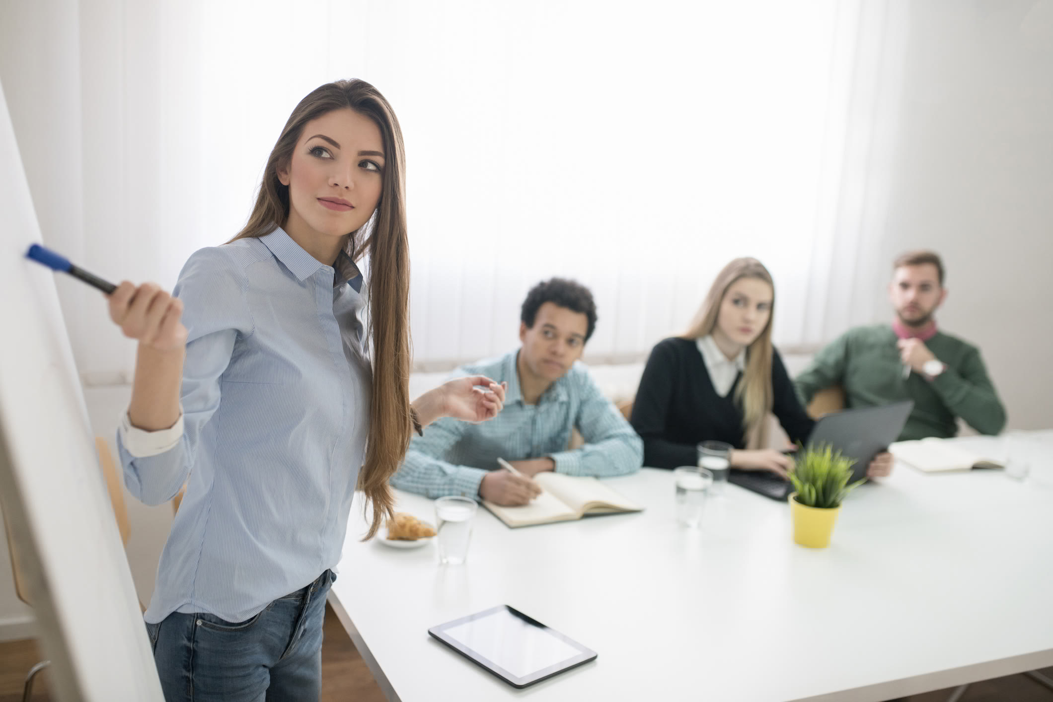 A woman stands at a whiteboard, holding a marker and looking to the side, while three people sit at a table with notebooks, a laptop, and glasses of water, appearing attentive during a meeting or presentation.