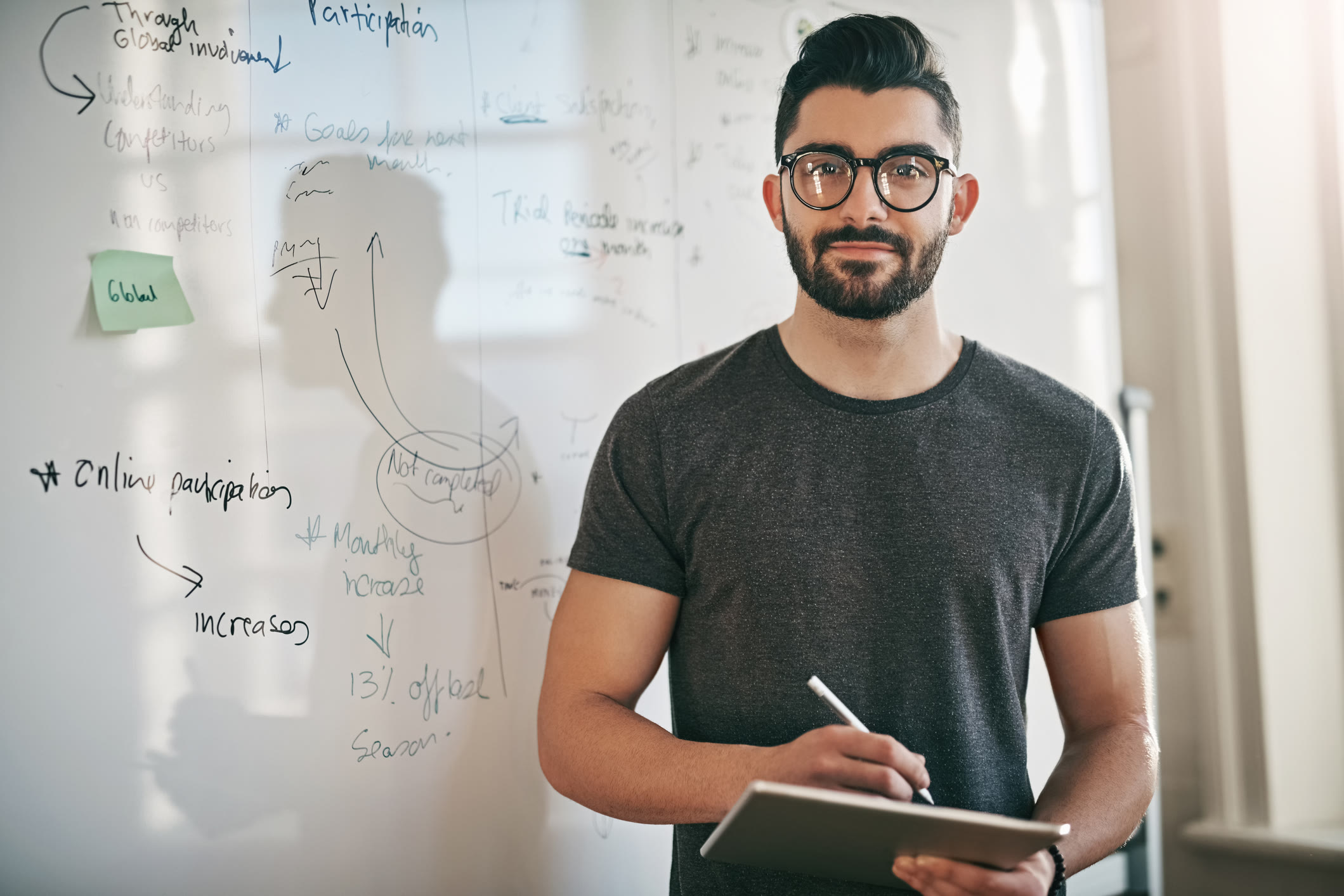 A man with glasses and a beard stands in front of a whiteboard covered in notes and diagrams, holding a digital tablet and stylus, looking confidently at the camera.